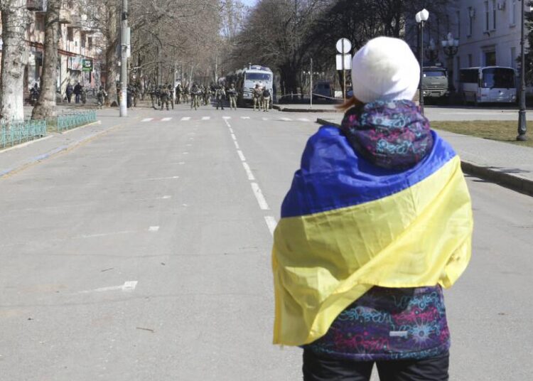 Seorang wanita yang ditutupi oleh bendera Ukraina berdiri di depan pasukan Rusia di sebuah jalan selama demonstrasi menentang pendudukan Rusia di Kherson, Ukraina, pada 19 Maret 2022. Menurut TV pemerintah Rusia, masa depan wilayah Ukraina yang diduduki oleh pasukan Moskow adalah segalanya. tetapi memutuskan: Referendum untuk menjadi bagian dari Rusia akan segera berlangsung di sana, dan penduduk yang gembira yang ditinggalkan oleh Kyiv akan dapat makmur dalam damai. Pada kenyataannya, Kremlin tampaknya tidak terburu-buru untuk menyegel kesepakatan di wilayah selatan Ukraina Kherson dan Zaporizhzhia dan provinsi timur Donetsk dan Luhansk.