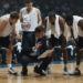 A basketball coach gives strategic instructions to the players during a timeout in the middle of a game. The players, with towels around their necks and leaning forward, listen intently as the coach points to the strategy board. A serious atmosphere is evident on the faces of the entire team as they focus on the explanation of game tactics.