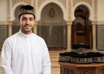 Pria muslim mengenakan baju koko krem dan peci hitam di halaman masjid.