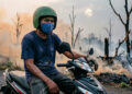 seorang pengemudi ojek online (ojol) berusia paruh baya duduk di atas sepeda motornya, mengenakan helm hijau tua dan masker kain berwarna biru
