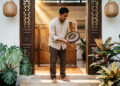 Seorang pria Indonesia berdiri di bingkai pintu utama rumahnya, memegang kompas feng shui (luopan) setinggi pinggang untuk mengukur arah hadap rumah. FOTO/AI for HAI.